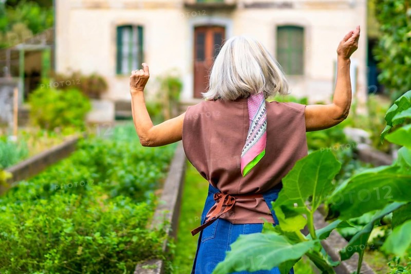 Senior woman with gray hair and a colorful scarf enjoying a sunny day outdoors, raising her arms in a carefree gesture in a lush green vegetable garden, embodying freedom and joy