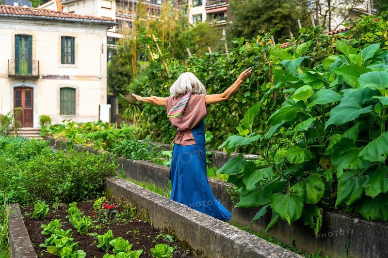 Senior woman with gray hair and scarf feeling freedom and happiness, walking with arms outstretched along a garden path with raised beds filled with plants