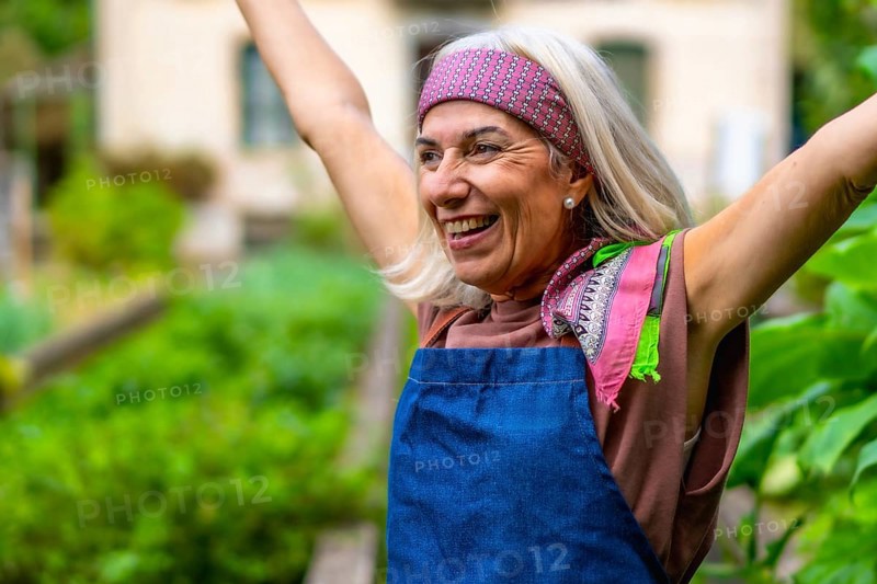 Senior woman in a bandana and apron raises her arms in a sunlit garden, smiling with joy and vitality as she celebrates healthy, active retirement and a flourishing outdoor hobby