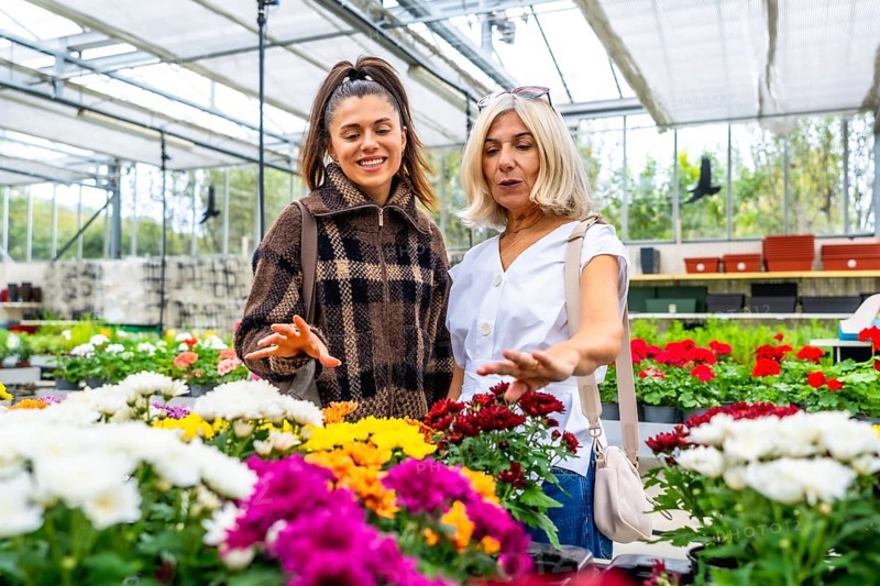 Mother and daughter smiling as they choose colorful potted flowering plants together in a bright greenhouse, enjoying a shared shopping and gardening outing