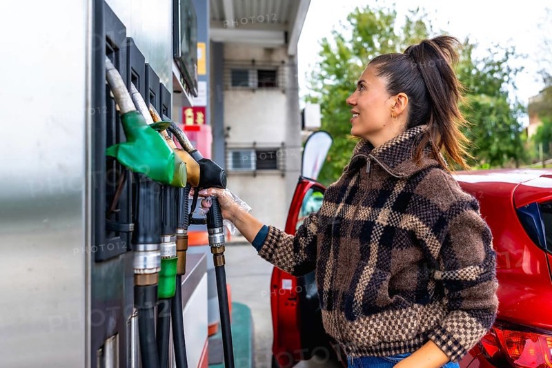 Woman in a casual plaid jacket holding a fuel pump nozzle, refueling her red car at a gas station, symbolizing transportation, energy consumption, and daily life