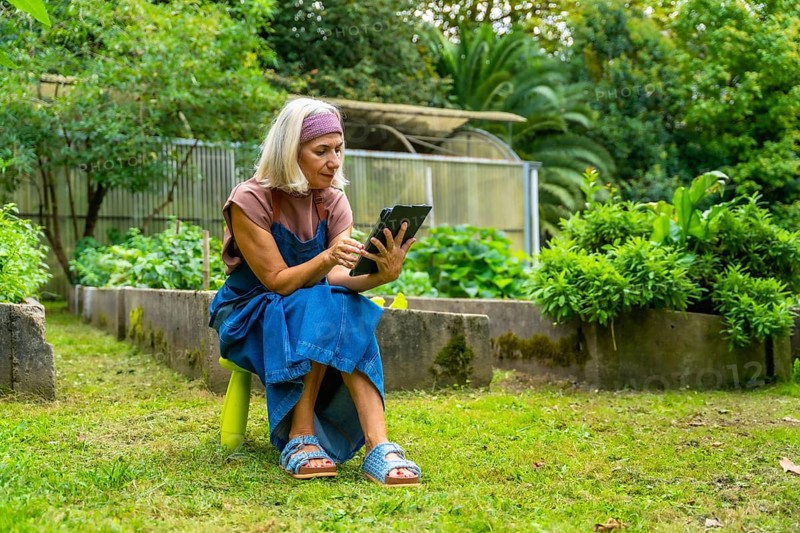 Senior woman sitting on a stool in her green garden, using a digital tablet for managing her plants and relaxing outdoors in nature with modern technology