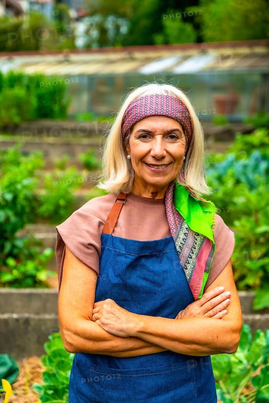 Senior woman wearing an apron and headscarf, standing with arms crossed in a lush community vegetable garden, smiling and looking directly at the viewer