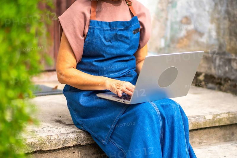 Senior woman in a blue apron working on a laptop outdoors, managing her small business and staying connected through remote work, technology and everyday entrepreneurship