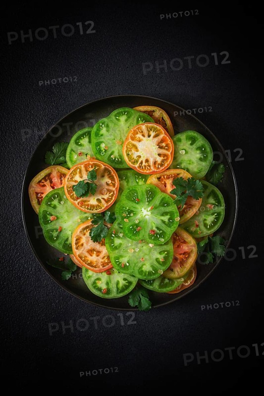 Sliced green tomatoes, on a dark surface, garnished with herbs and salt, top view
