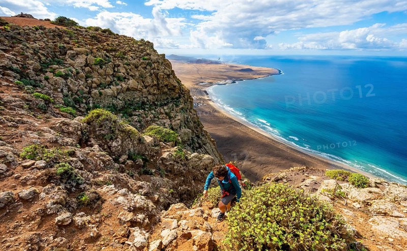 Female hiker climbing to Castillejo summit, view from the Risco de Famara cliffs of the coast and the sea with Famara beach, Playa de Famara with La Calaeta, Lanzarote, Canary Islands, Spain