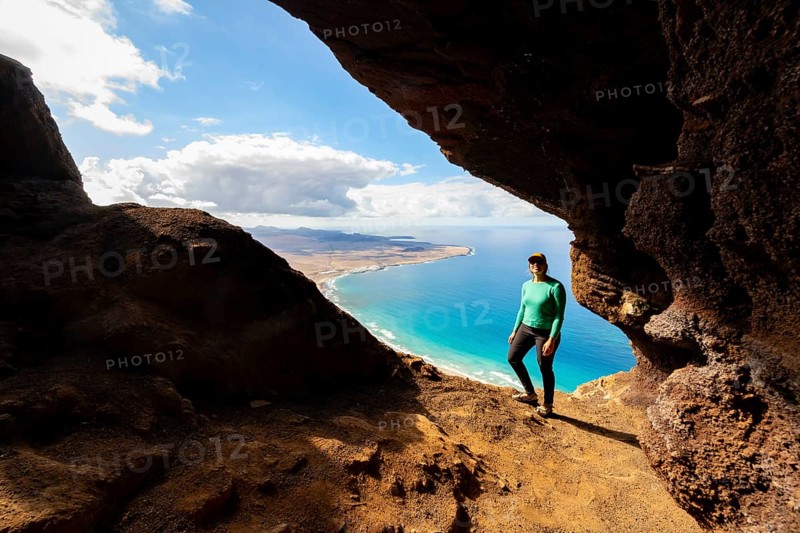 Tourist in the Cueva de las Cabras cave, young woman enjoying the view from the Risco de Famara cliff to Famara beach, Playa de Famara with La Calaeta, Lanzarote, Canary Islands, Spain