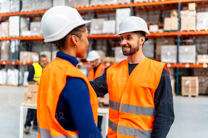 Two diverse warehouse workers wearing safety hard hats and vests are confidently smiling and collaborating in a logistics distribution center, demonstrating teamwork and professional communication