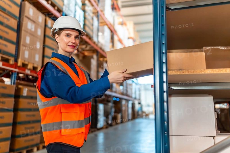 Woman worker wearing safety helmet and reflective vest, putting a cardboard box on a shelf in a large logistics warehouse, managing inventory and shipping operations
