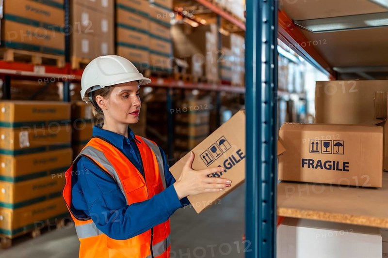 Worker in safety gear handling a fragile package, organizing inventory on a shelf in a modern distribution warehouse, ensuring efficient supply chain management and storage