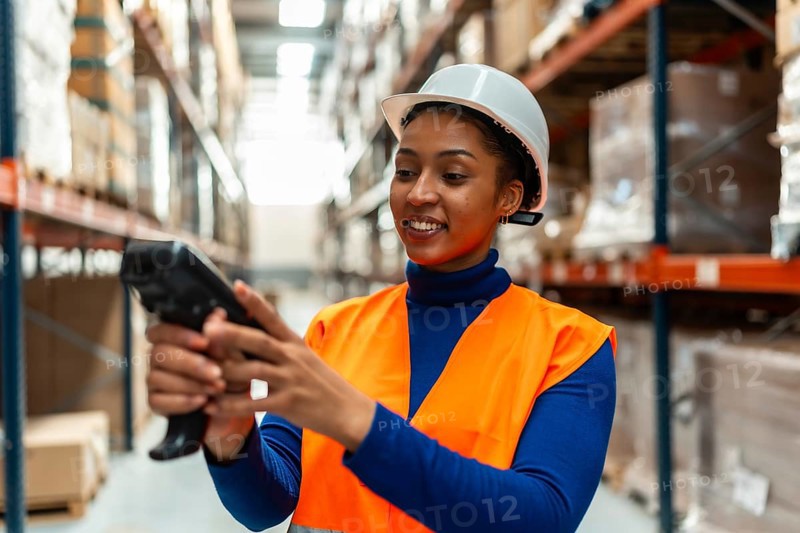 Young woman warehouse worker in safety hard hat and reflective vest scanning inventory with a barcode scanner amid racks of boxes, smiling while managing modern logistics operations