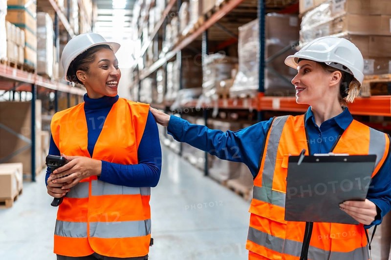 Two diverse women warehouse workers in safety vests and hard hats smiling and collaborating, discussing inventory and operations in a modern logistics environment