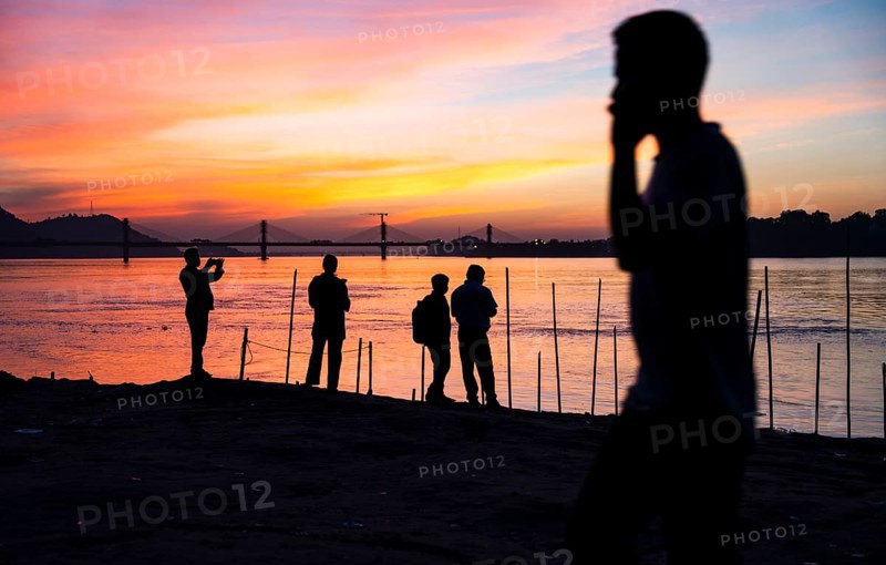 People enjoy the vibrant sunset by the Brahmaputra riverside, capturing photos and moments against the scenic backdrop of a bridge and glowing sky, on November 4, 2025 in Guwahati, India