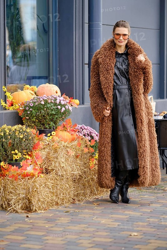 A woman stands next to the autumn installation, wearing a brown coat and black dress. She poses next to autumn decorations, including pumpkins and flowers, highlighting the festive atmosphere