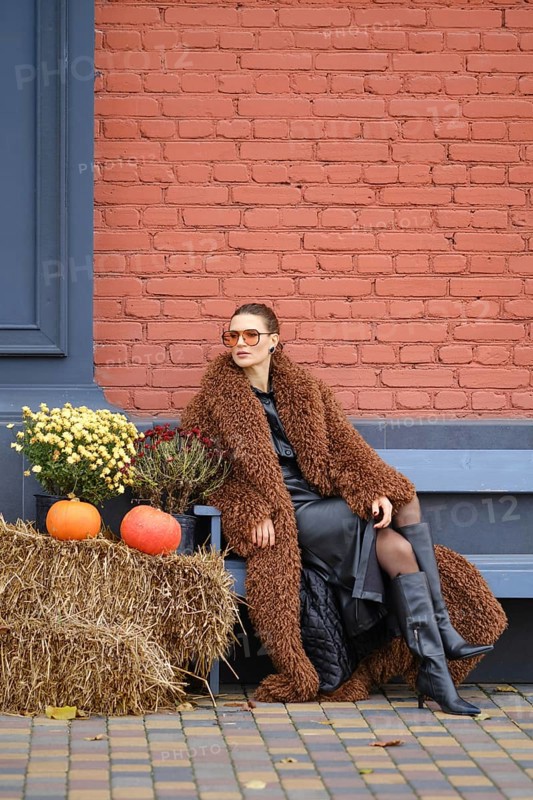 A woman sits on a bench dressed in warm fur coat. She poses near colorful pumpkins and vibrant flowers against a red brick wall, embodying the autumn spirit