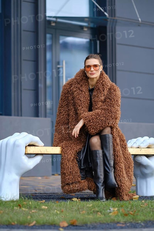 A stylish woman wearing a brown fur coat and black knee-high boots poses on a unique bronze bench supported by large sculpted hands. The background features a modern building
