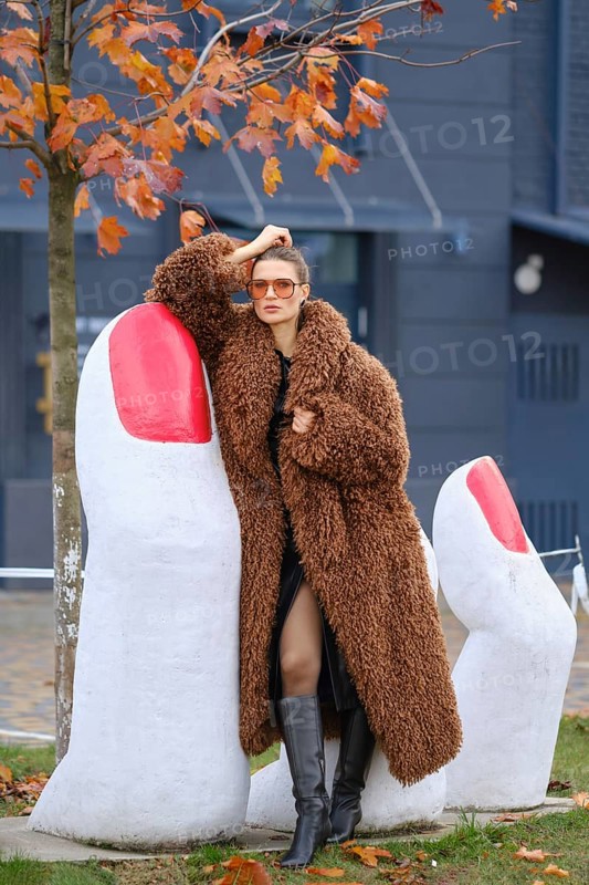 A fashionable woman in a cozy brown coat stands near large white sculptures resembling fingers with red nails. Colorful autumn leaves surround her as she strikes a confident pose outdoors