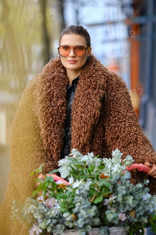 A woman in oversized sunglasses and a fluffy brown coat strolls through a vibrant urban area. She is pushing a cart filled with lush green plants and colourful flowers on a sunny day