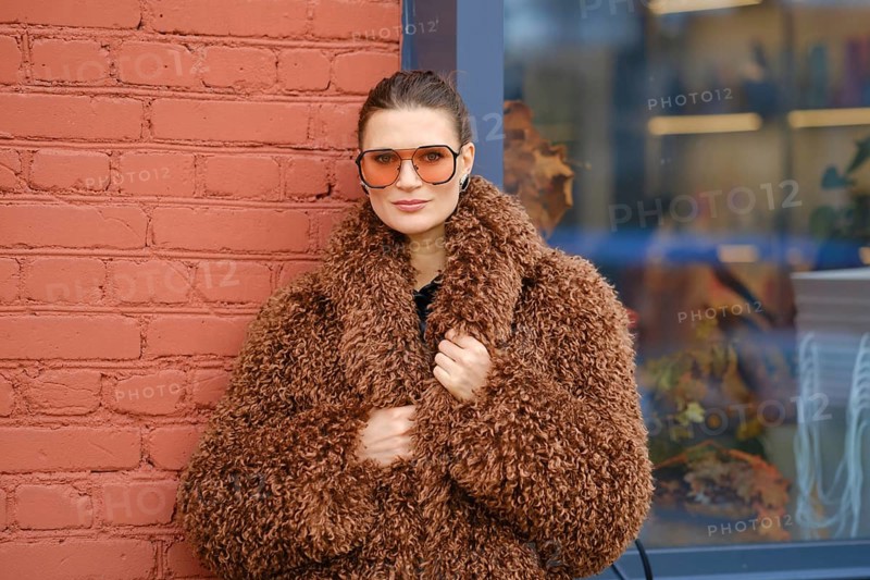 A woman poses confidently in a warm, brown, fluffy coat and oversized sunglasses. She stands against a red brick wall, showcasing a modern urban style in a vibrant setting