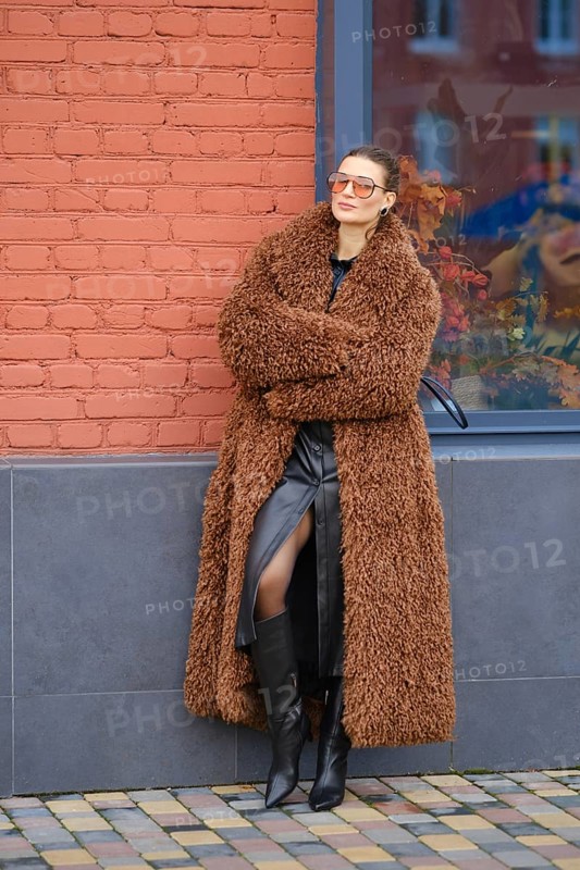 A fashion model stands with crossed arms beside a red brick wall. She wears a fluffy brown coat, a black dress, and knee-high black boots, appearing stylish and relaxed in a vibrant urban setting
