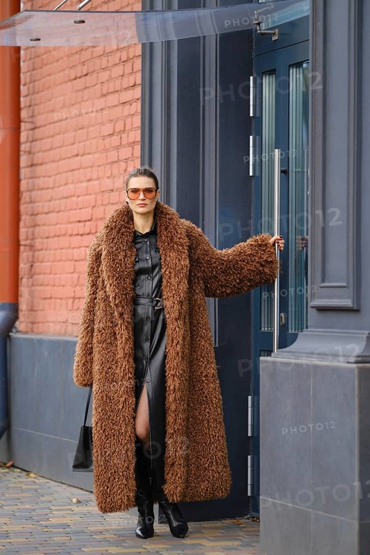 A fashionable woman in a long brown fur coat and sunglasses walks out of a colorful urban office building