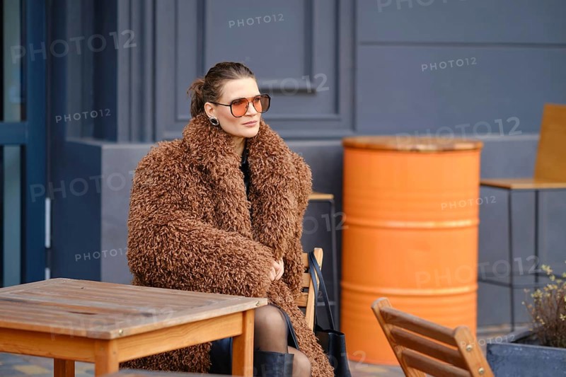 A fashionable woman sits outside a cafe, wearing a large brown fur coat and stylish sunglasses. She appears relaxed as she enjoys the atmosphere in an urban environment during daylight