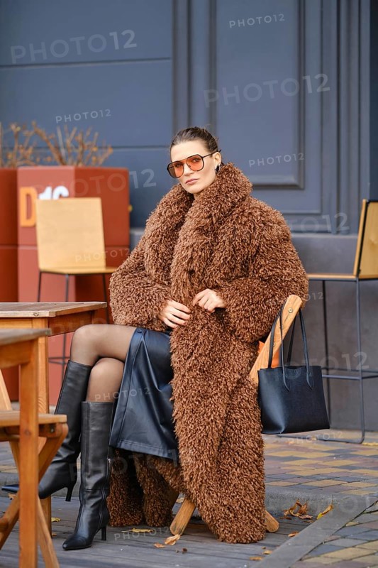 A stylish woman sits at an outdoor cafe during autumn, wearing a fuzzy brown coat and black knee-high boots. She pairs her look with oversized sunglasses and handbag