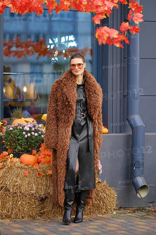 A woman dressed in a stylish brown fuzzy coat and black leather dress stands next to a seasonal display of pumpkins and autumn leaves