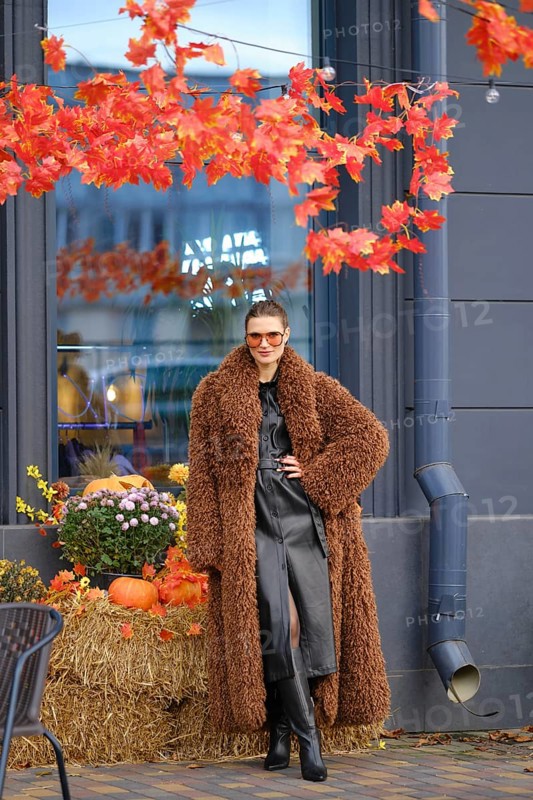 A stylish model stands near a store with bright autumn leaves overhead. She wears a brown fur coat and sunglasses. Pumpkins and flowers add seasonal charm to the cozy setting
