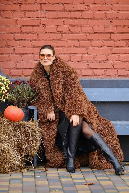 A woman wearing a brown fuzzy coat and knee-high black boots poses confidently outdoors. She is surrounded by autumn decorations, including a pumpkin, haystack and flowers, set against a brick wall