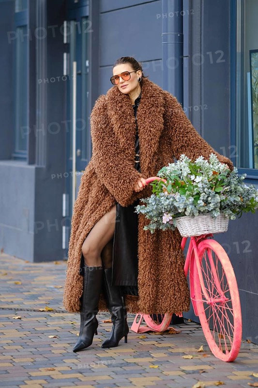 A fashionable woman stands against a modern building, wearing a cozy brown coat and tall black boots. She leans on a pink decorative bicycle adorned with fresh flowers, enjoying her city surroundings