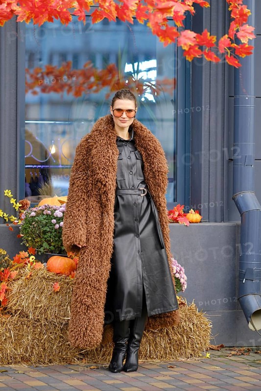 A stylish woman poses outside a farmer's grocery shop decorated for autumn, wearing a brown fur coat and sunglasses. Colorful pumpkins and flowers add to the seasonal charm