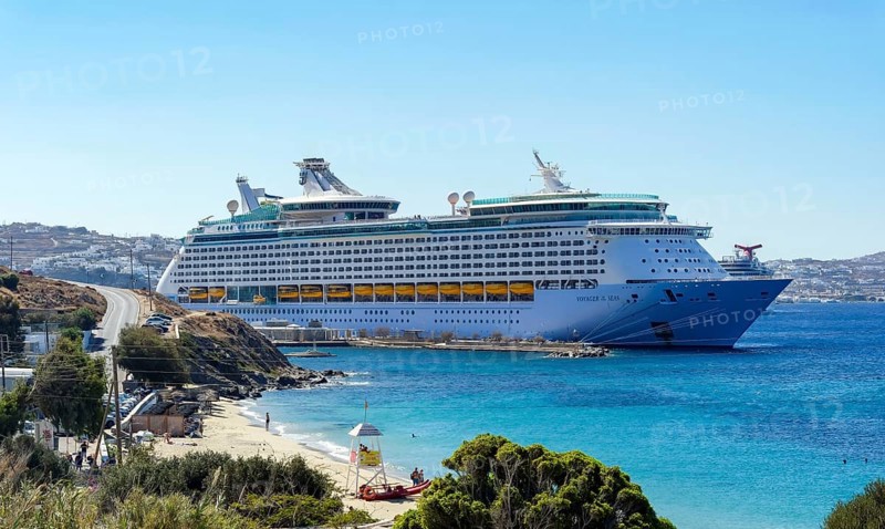 Mykonos, Cyclades, Greece - Voyager of the Seas cruise ship is located in Mykonos Harbour Bay. In front, tourists bathe on the sandy beach of Paralia Agios Stefanos. In the back is the old town of Mykonos
