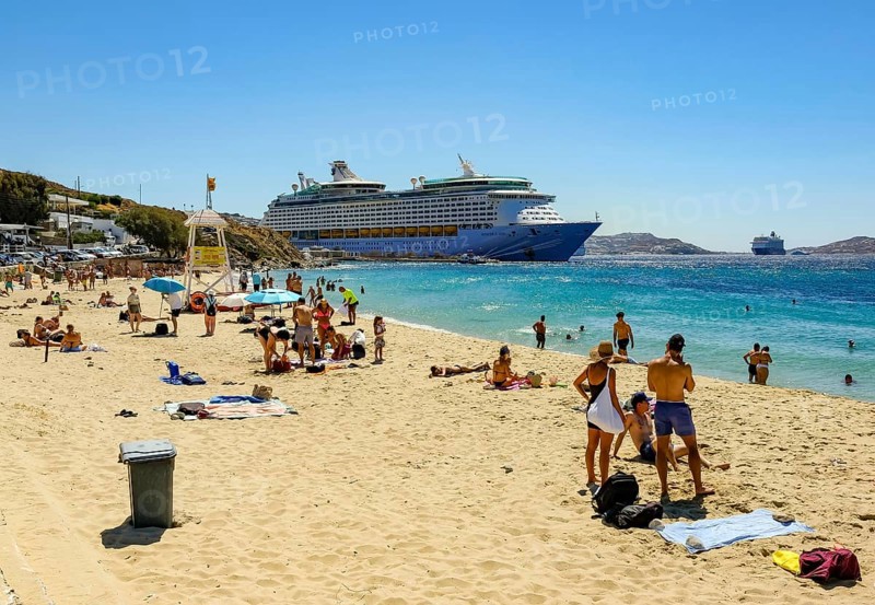 Mykonos, Cyclades, Greece - Voyager of the Seas cruise ship is located in Mykonos Harbour Bay. In front, tourists and locals bathe on the sandy beach of Paralia Agios Stefanos