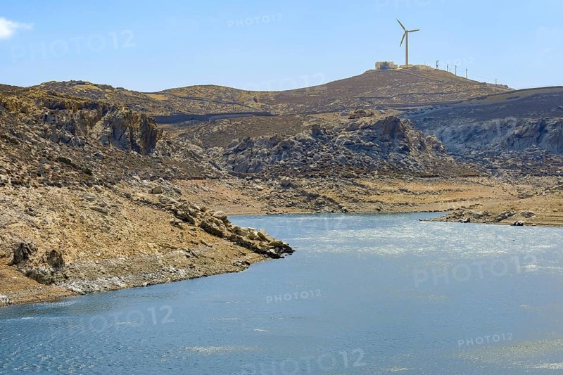 Mykonos, Cyclades, Greece - Dry Fokos Reservoir near Ano Mera. The Mykonos Municipal Water Supply and Sewage Management Company has two dams, Marathi Reservoir and Fokos Reservoir, to supply water to the island. Due to the prolonged drought, both are almost empty and are not being used. A wind turbine at the back of the mountain