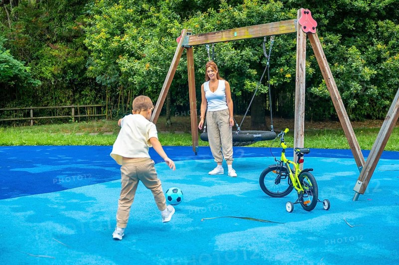 Young boy kicking a soccer ball on a playground while mother watches, with a swing set and bicycle nearby, creating a joyful scene of family fun on a sunny day