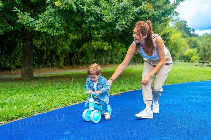 Young mother helping her son learn to ride a balance bike on a blue surface in a green park, promoting childhood development and family bonding