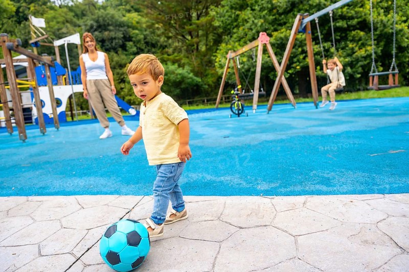 Happy toddler kicking a vibrant soccer ball at the playground, with mother and sister enjoying playtime in the background, creating a joyful scene of family togetherness and fun