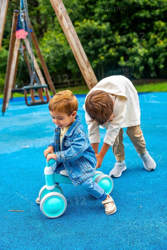Older brother is helping his younger brother to ride a balance bike on a blue playground surface with a swing set in the background, promoting childhood fun, learning, and brotherly bonding