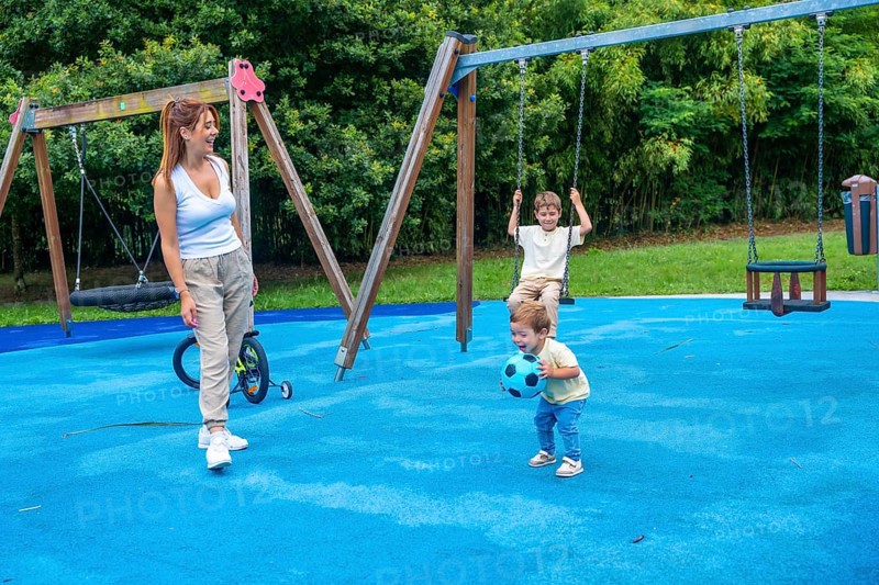 Mother enjoying a sunny day at the playground, watching her children happily playing with a ball and swinging on the swings, creating joyful moments of childhood and togetherness