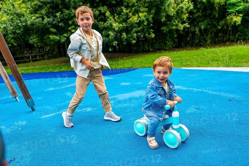Two brothers are spending time together on a playground, one riding a balance bike and the other walking next to him on a blue rubber surface