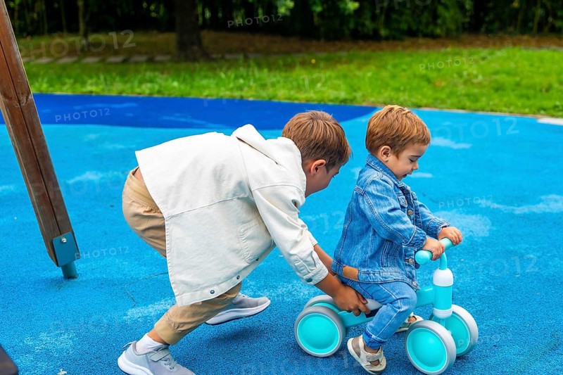 In a vibrant playground setting, an older brother supports his younger sibling as he learns to ride a balance bike, fostering a sense of camaraderie and shared joy in outdoor play