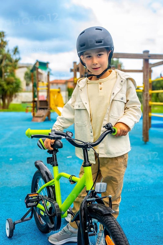 Young boy wearing a helmet standing and holding the handlebars of his bicycle with training wheels in a playground with a blue rubberized floor