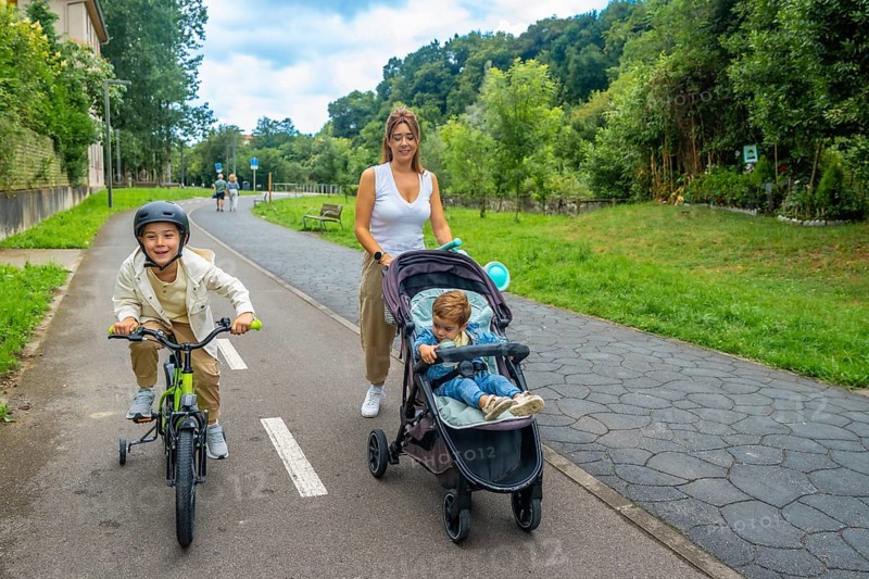Mother pushing a stroller while walking alongside her son riding a bicycle in a vibrant park, cherishing quality time together outdoors and enjoying the summer sunshine