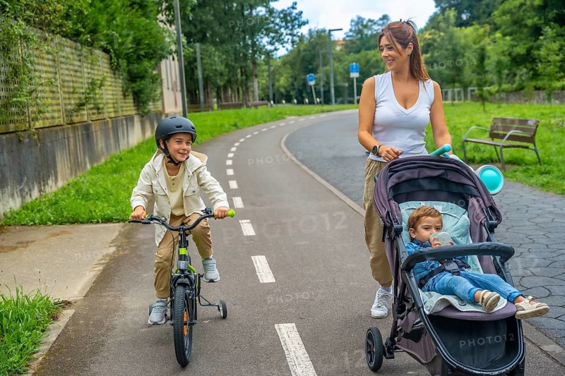 Happy mother walking with her two children, one riding a bicycle with training wheels and the other sitting in a stroller, enjoying a sunny day at the park