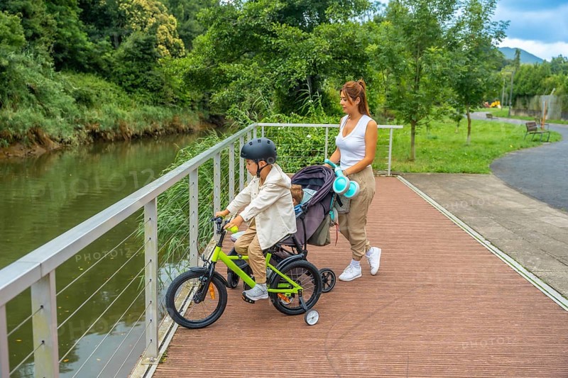 Young mother pushing a stroller with a baby inside and watching her other son riding a bicycle on a wooden walkway next to a river in a park during a sunny summer day