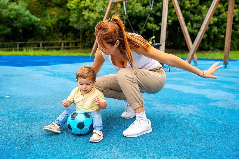Young mother crouching beside her toddler son, guiding him as he plays with a soccer ball on a vibrant blue playground surface, nurturing early love for sports