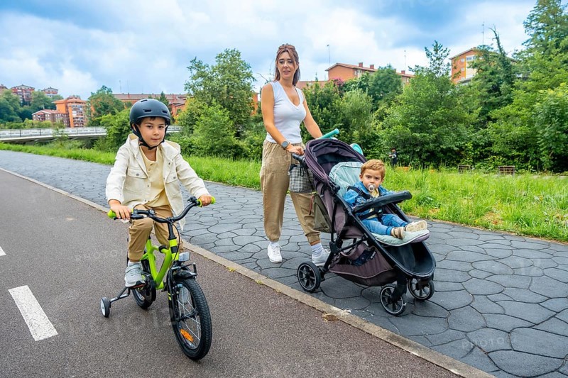 Young mother pushing a stroller with her baby boy, while her older son rides a bicycle with a helmet, enjoying a sunny day filled with laughter and togetherness in the park