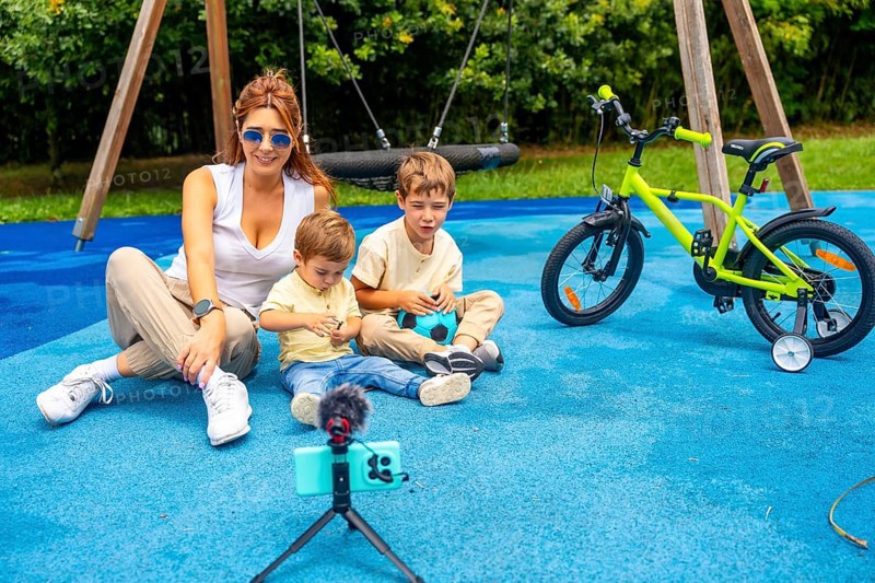 Influencer mother recording a video with her two sons at playground using a smartphone and microphone on a tripod, with a kids bicycle in the background