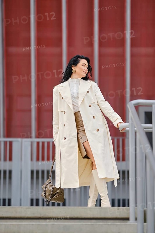 A woman elegantly poses on a staircase in a stylish autumn outfit featuring a long coat and knee-high boots. The vibrant red building creates a striking backdrop for her modern look